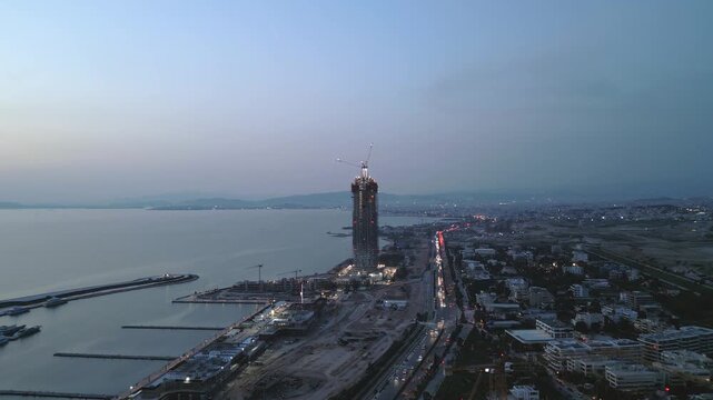 Aerial drone footage focused on a skyscraper under construction during blue hour. High-rise tower development with cranes and structural framework rising above a large urban redevelopment area