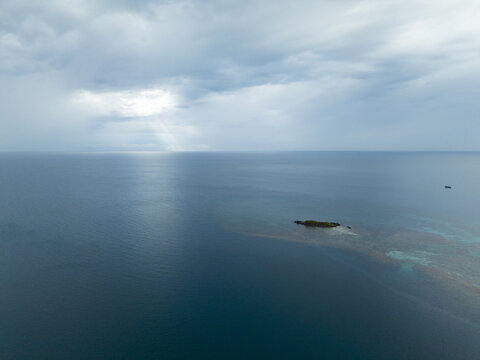 Aerial view of an islet surrounded by turquoise shallows amidst the deep blue sea under a sky streaked with sunlight, Karimunjawa, Central Java, Indonesia.