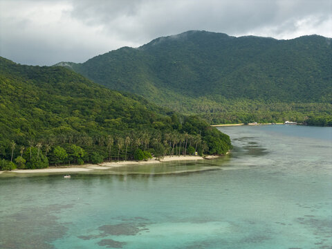 Aerial view of the turquoise waters gently lap against the pristine shores, embraced by lush green hills under a cloudy sky, Karimunjawa, Central Java, Indonesia.
