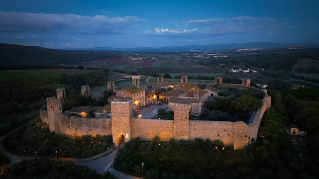 Aerial view of a medieval walled town glows warmly against the deep blue dusk, its ancient stones whispering tales of centuries past, Monteriggioni, Tuscany, Italy.