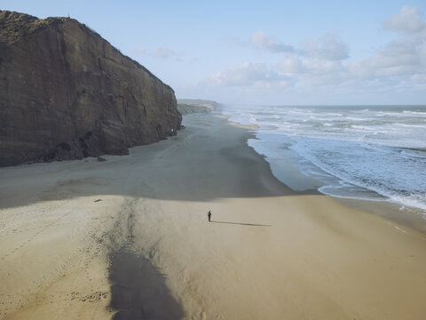 Aerial view of a solitary figure stands dwarfed by the vast expanse of the sandy beach, where the rugged cliffs meet the turbulent sea, Lagoa de Obidos, Leiria District, Portugal.