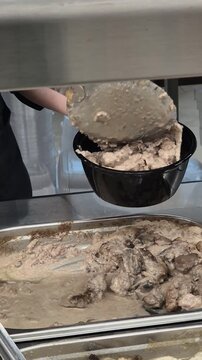Stew serving cafeteria worker scooping creamy meat and mushroom dish into a black plastic container at a commercial food service station