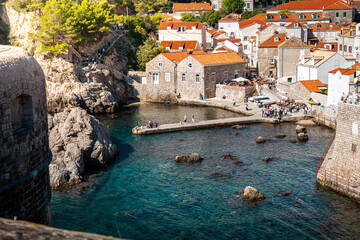 landscape shot of harbour beach on Adriatic Sea in dubrovnik old town croatia with red roof houses and clear blue water with tourists walking around and bright green trees in summer with sunlight  © STJMEDIA