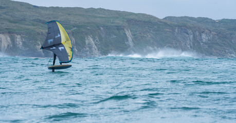 Naklejka premium A man wing foils at sea using a yellow and grey hand held inflatable wing, riding a hydrofoil surfboard. Green sea, cloudy blue sky. Coastal cliffs in the background.