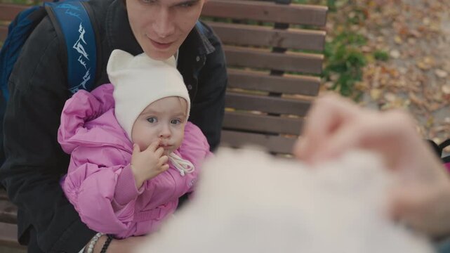 Family baby on bench reaches snack. Autumn park scene with toddler in pink puffer and white knit hat, caregiver offering treat while parent holds child, stroller beside them, warm tender bonding
