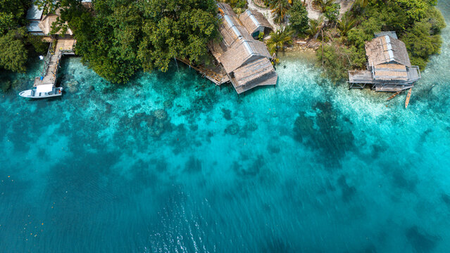Aerial view of vibrant turquoise waters meet the lush green shoreline dotted with rustic huts and a wooden pier, Solomon Islands, Solomon Islands.