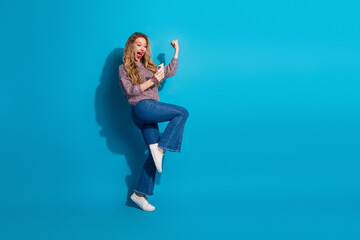 Young woman jumps with phone in hand exuding joy fashion and energy against blue background