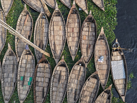 Aerial view of wooden boats clustered tightly together on the river's surface, their textures contrasting with the patches of green vegetation, Dhaka, Dhaka Division, Bangladesh.