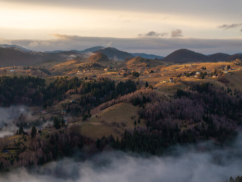 Aerial view of a serene landscape where fog gently hugs the undulating hills and the distant mountains fade into the soft glow of the setting sun, Sirnea, Brasov, Romania.