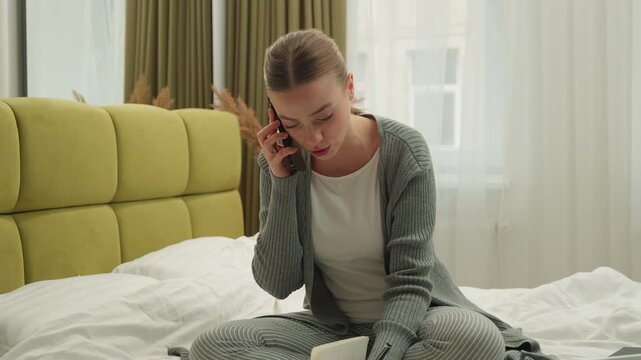 Multitasking stressful Caucasian woman talking mobile phone writing notes using laptop at home bedroom on bed communicate smartphone call write computer browsing distant learning freelance multitask