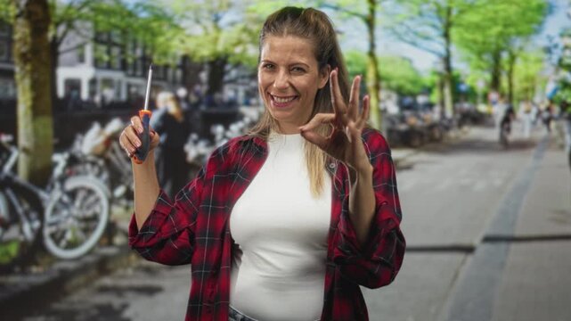 Woman wearing red plaid shirt holding a screwdriver and making ok sign with hand on a busy street with bicycles and trees; confidence repair.