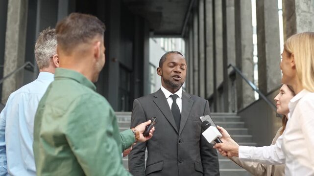 Inquisitive multiethnic journalists with microphones talking to famous businessman or politician near office on street. Correspondents interviewing successful man. Breaking news.