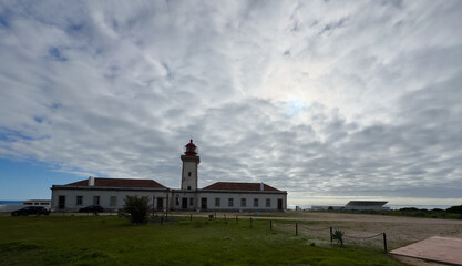 Fototapeta premium Alfanzina Lighthouse with red lantern tower rising above green coastal vegetation and rocky landscape.