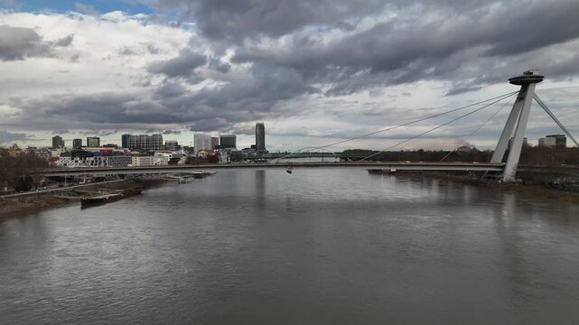 Aerial view of the iconic SNP Bridge stretching over the calm, grey Danube river under a cloudy sky, Bratislava, Bratislava Region, Slovakia.