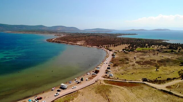 Aerial view of a beach with turquoise waters meeting the sandy shore, cars parked along the road, contrasting with the dry landscape, Cunda Island, Ayvalik, Balikesir, Turkey.