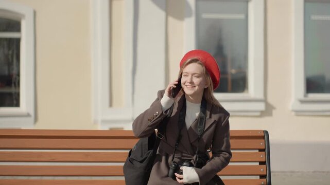 Young caucasian woman by bench making a thoughtful phone call in soft sunlight brown coat, red beret, tote bag on shoulder, city facade backdrop, alternating walking and seated frames show commuter,