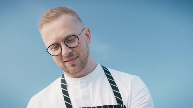 Caucasian male chef chopping red bell pepper on rooftop table with fresh ingredients closeup. Man in apron preparing food outdoors with urban cityscape in background. Cooking session under sunny sky