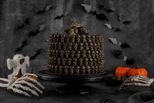 Halloween chocolate cake decorated with chocolate skulls and a skeleton hand on a black cake stand. Spooky ghost, pumpkin, and bat decorations create a dark festive atmosphere.