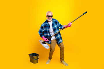 Happy senior man cleaning with mop and bucket on bright yellow background, wearing sunglasses,...