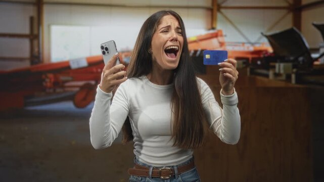 Woman holding smartphone and creditcard in hand, mouth open shouting while checking device inside a building; financial frustration.