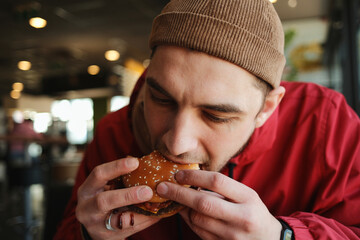 Close up of a young man biting a juicy beef burger with bacon on a sesame bun while sitting in a fast food restaurant. Concept of casual dining, street food and quick urban meal © Ekaterina