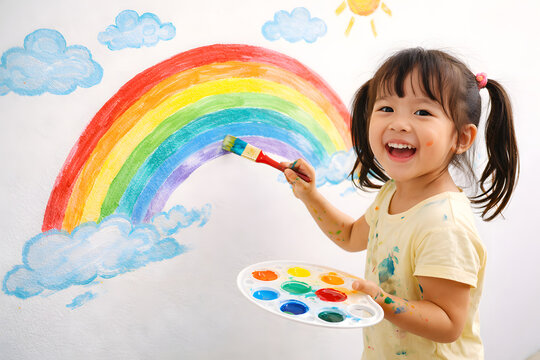 Child painting rainbow on wall, happy little girl holding paintbrush and palette, creative kid drawing colorful arc with clouds and sun, preschool art activity, early childhood education creativity, p