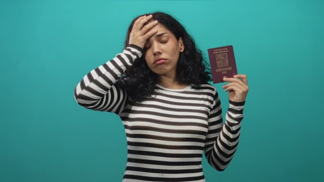 Young brunette woman holding czech passport with hand on forehead in studio against teal backdrop; travel frustration.