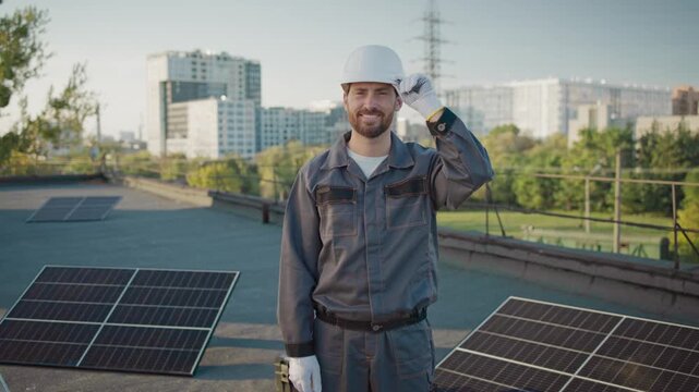 Caucasian male engineer in helmet and gloves standing next to solar panel on rooftop. Confident worker smiling to camera at solar installation. Urban cityscape in background. Ecofriendly energy setup