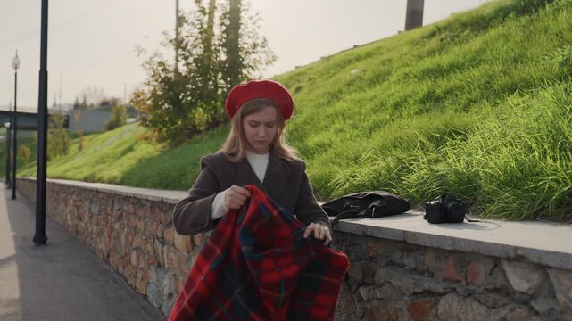 Caucasian woman folding red plaid scarf on harbor promenade near grassy slope, wearing red beret and brown coat, hands smoothing wool fabric over stone wall, vintage camera and bag nearby, closeup