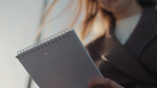 Woman holding spiral notebook closeup, reporter jotting notes during interview on city street, warm backlight rim highlighting hair, shallow depth of field, professional coat, subtle hand movements,