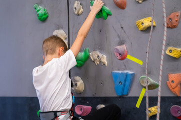 Fototapeta premium Young Boy Climbing an Indoor Bouldering Wall with Colorful Holds at Sport Gym Wearing a Harness