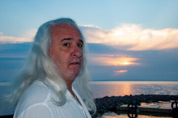 Fototapeta premium Senior lifestyle. Natural aging beauty. Mature male grooming. Mature man by ocean horizon during sunset. Healthy long hair at older age. Authentic masculine style. Retired man enjoying evening