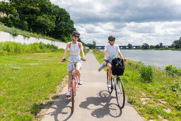 Fototapeta premium Women riding bicycles along riverside path during summer day with green park landscape and cloudy sky. Active lifestyle, friendship, outdoor recreation healthy activity travel freedom leisure cycling