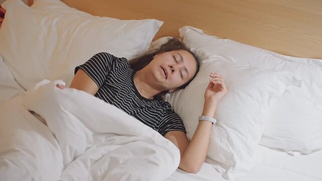 Hotel bed guest lying exhausted under white linen, polkadot shirt, wristband on hand, soft pillow and plush mattress, warm wood headboard, quiet evening atmosphere, slow breathing, peaceful surrender