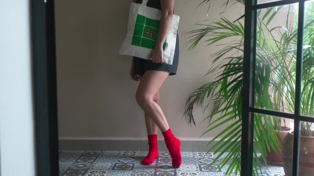 Young woman standing indoors near a window wearing red socks and holding a reusable artistic tote bag. Natural light, indoor plants and patterned floor create a calm modern lifestyle atmosphere.