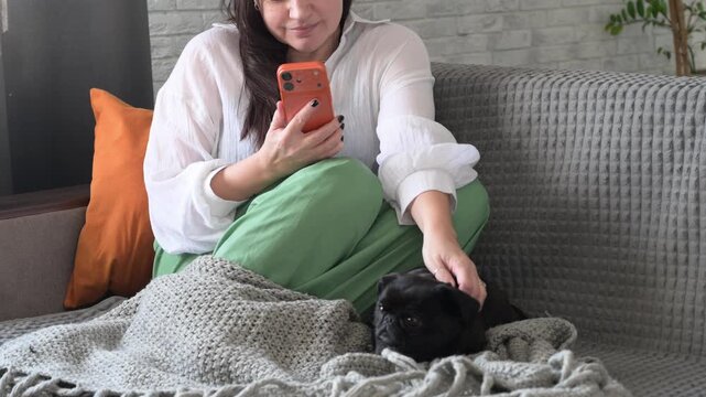 Woman sitting on sofa drinking coffee, using smartphone, and petting griffon dog. Concept of relaxation, anti stress lifestyle, cozy home atmosphere, and living with pets.
