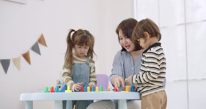 Asian female preschool teacher coaching toddler boy, toddler girl building colorful peg stack tower on table, playful classroom bunting decor, early learning routine, slow motion education content.