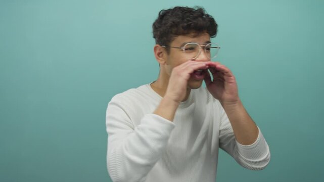 Young hispanic man wearing glasses cups hands to mouth and opens mouth to shout in studio with teal backdrop; urgent alert.