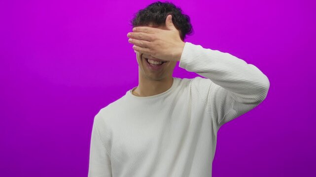 Man covering eyes with hand in purple studio setting, wearing ribbed white sweater and wireframe glasses with a slight smile; shyness.