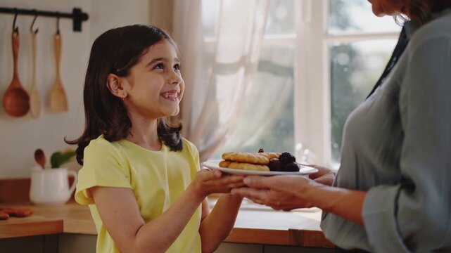 Handing mother in blue shirt passing pancakes with berries in kitchen as child reaching for meal