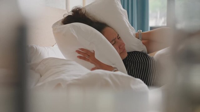 Exhausted hotel guest smothers sound with pillow while lying by window side view through blurred foreground reveals woman in polka outfit covering ears with fluffy pillow, teal curtains and bedside