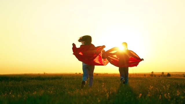 Kids girls play superheroes in red capes in park on green grass, dream. Children in red capes run across meadow sunset. Happy children run play superheroes in nature. Children in capes play heroes