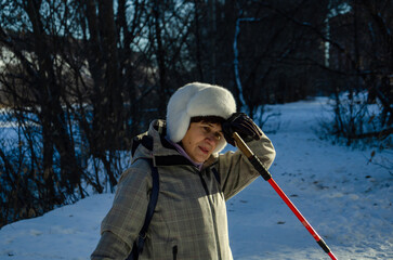 Senior woman in white fur hat resting during Nordic walking in snowy winter forest. Leaning on red pole, wiping brow. Winter fitness break, active aging, outdoor exercise