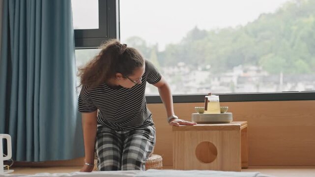 Woman hotel window turning toward view, student guest exploring ledge and settling to gaze at city panorama casual plaid pants, light filtering through curtains, small wooden tray on side table,