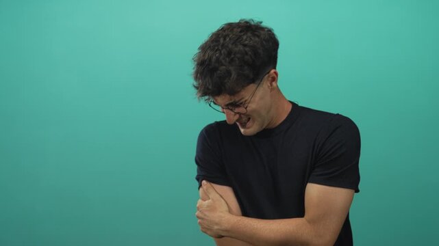 Man clutching upper arm and wincing in teal studio set, wearing glasses and black tshirt; pain vulnerability.