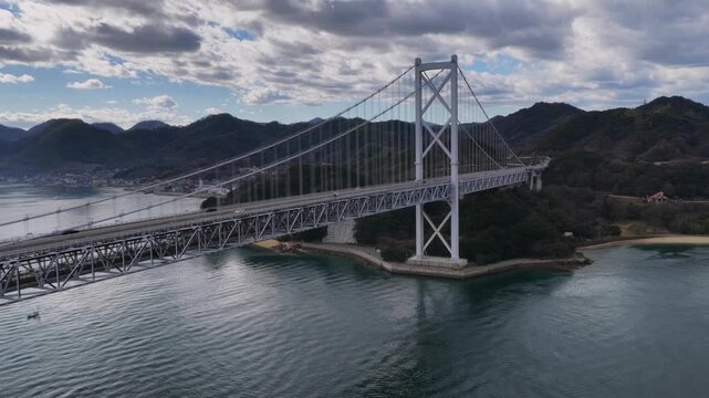 Cinematic aerial orbit of Innoshima Bridge under dramatic silver clouds, transition from backward pull to nose-in circle and forward flight.