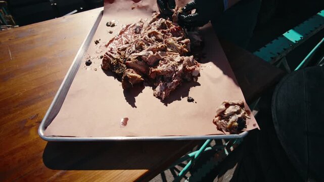 Black-gloved hands gather and consolidate shredded pulled pork on pink butcher paper in a metal tray in an overhead slow-motion shot