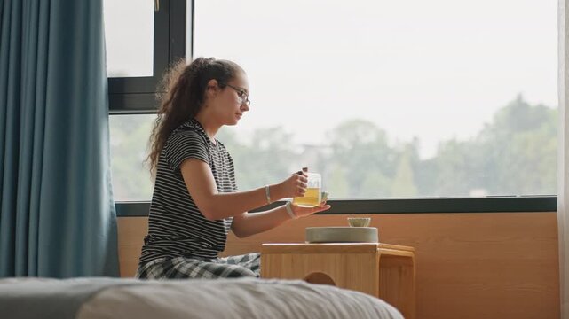 Hotel bedside breakfast girl pours juice from pitcher into glass at small wooden table next to bed, bright window view with treeline, soft curtains and hotel room bedding in foreground, playful