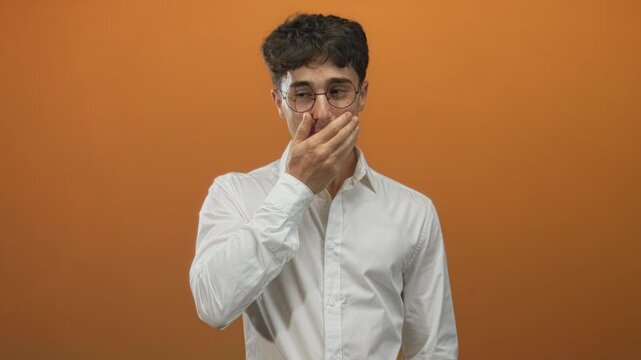 Man covering face with hand and glasses pushed up while wearing white shirt in orange studio, slight smirk visible; embarrassment doubt.