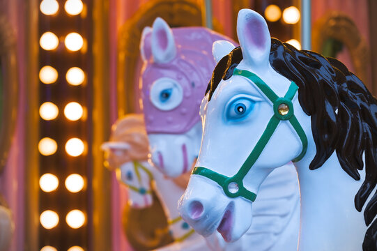 A white carousel horse with a black mane and blue eyes, wearing a green bridle, is in sharp focus against a blurred background of a pink and gold carousel with warm lights. 